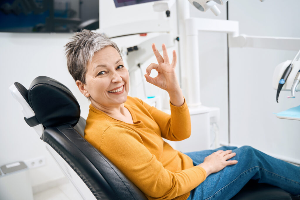 Smiling middle-aged lady giving the OK sign while sitting in a dental chair. 
